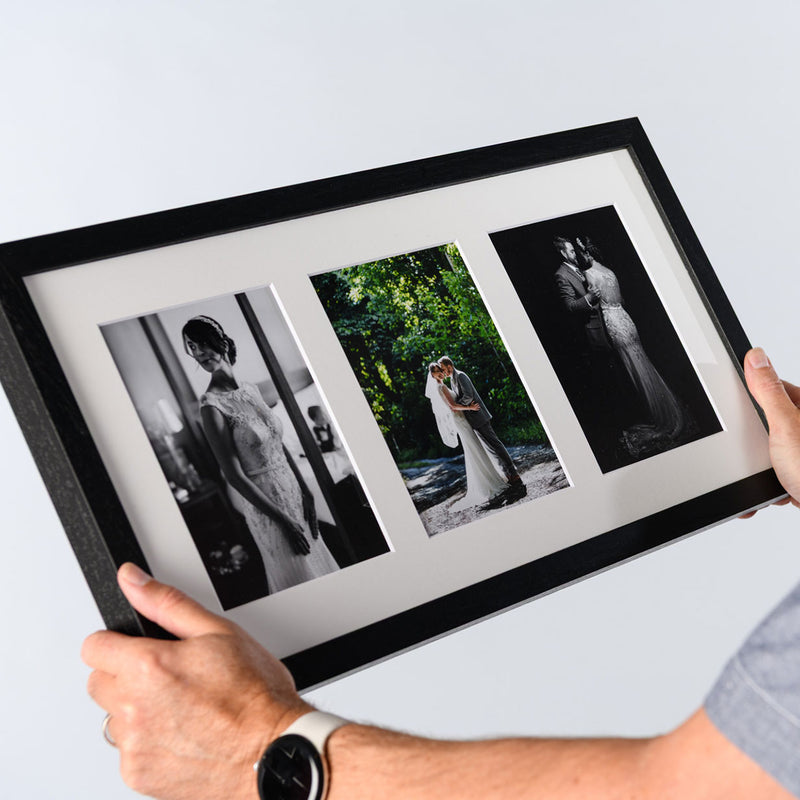 A person holds a black triple aperture multi photo framed wedding collage, including two black-and-white portraits and one color shot, against a light gray background. The frame is 50x25cm in size. 