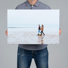 A man holding a 60x40cm canvas print featuring a same sex couple walking on the beach with their dog