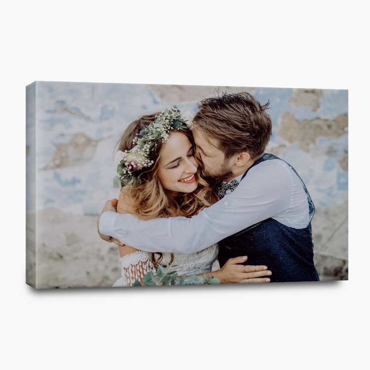 A rectangular canvas wall art print showing a couple embracing, displayed at an angle against a clean white background. The artwork features a smiling woman with a floral crown and a man in a white shirt and dark vest, set against a textured blue and beige wall.