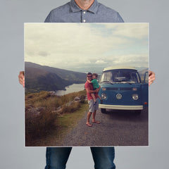 A person holds up a large square 70x70cm photo canvas print featuring a family scene with a blue Volkswagen van on a road by a lake.