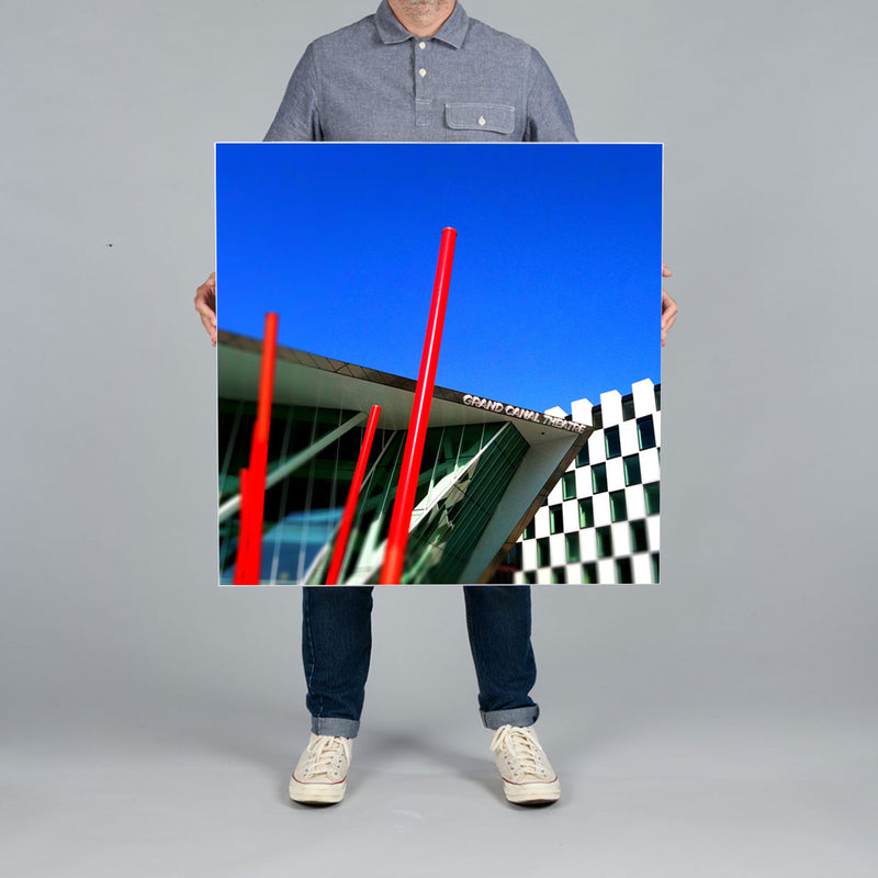 A person holds a square 80x80cm canvas print against a plain gray studio background. The canvas features a photograph of the Grand Canal Theatre in Dublin Docklands. 