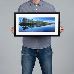 A person holds up a black-framed landscape print of a serene lake with pine trees and green hills under a blue sky, against a solid gray background.