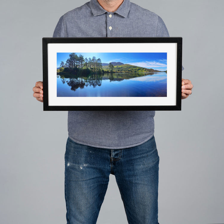 A person holds up a black-framed landscape print of a serene lake with pine trees and green hills under a blue sky, against a solid gray background.