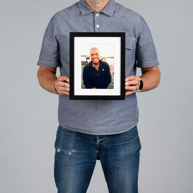 A man from The Canvas Works holds a 15x20cm black-framed custom portrait photo print against a solid gray background.