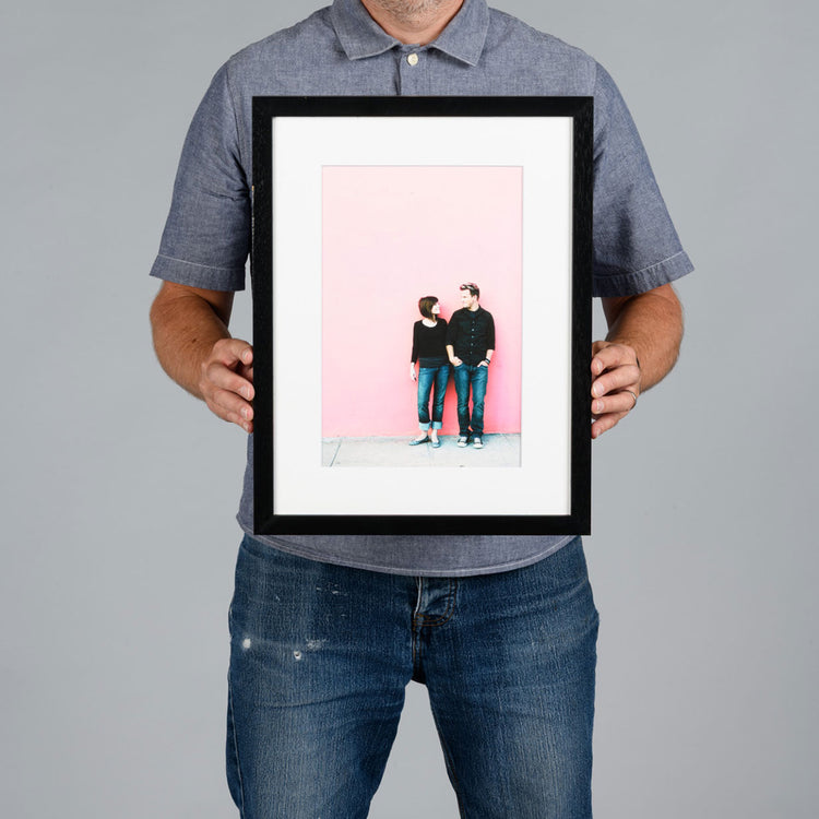 A man holding a 20x30cm framed photo print against a solid gray background. The print features a minimalist illustration of a couple standing against a solid pink background, enclosed in a white mat and a black frame.