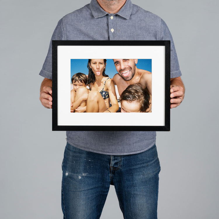 A man holds a framed 30x20cm custom photo print against a solid gray background. The black-framed print features a white mat and a colorful photograph of a family.