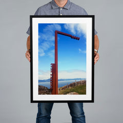 A man holds a large 40x60cm black framed photo print featuring a coastal landscape with a reddish-brown metal sign against a blue sky. 