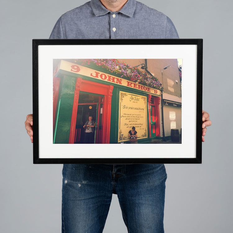 A person holding a 45x30cm white-framed art print of a colorful pub exterior with red and green facade details, set against a solid gray background. The pub is Kehoes in Dublin. 