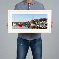 A person holds a white framed art print of a harbour scene in Kinsale with boats and masts against a plain gray background. The frame is white with a white matt. 