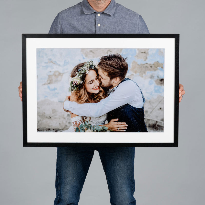 A man holds a large 60x40cm black-framed custom photo print featuring a wedding couple embracing. The print has a wide white mat border and is displayed against a solid gray background.