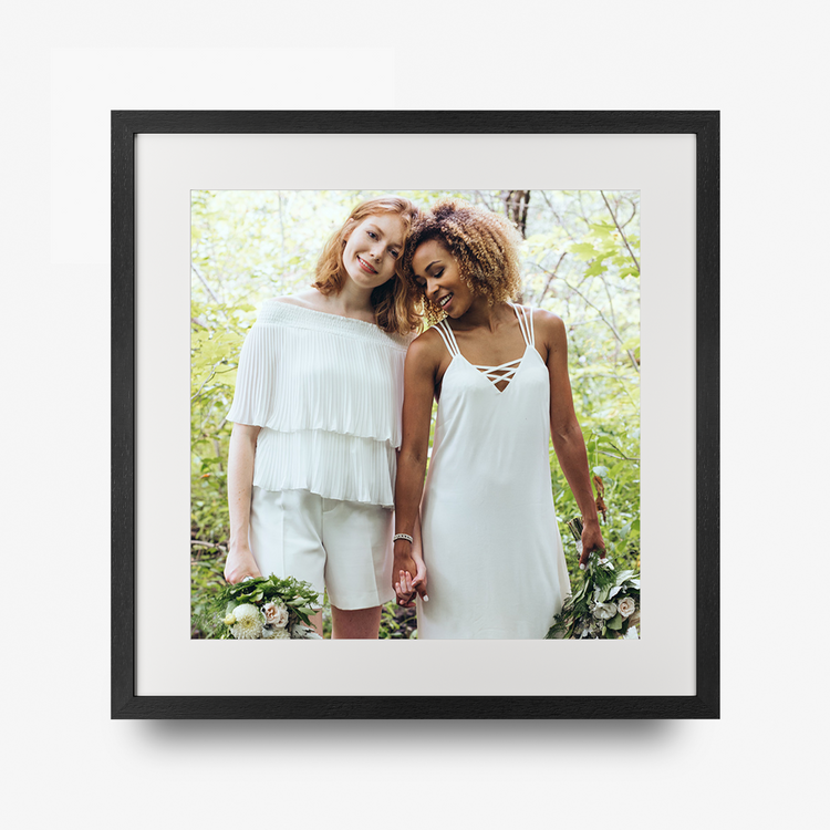A square black-framed custom photo print on a clean white background. The print features a photograph of two women in white outfits standing outdoors in a lush green forest, surrounded by a wide white mat and a sleek black frame.